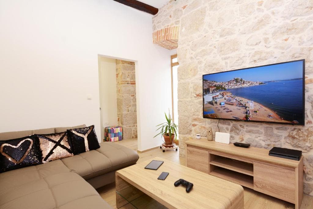 Living room in Design Apartment X with a beige corner sofa, a wooden coffee table and a wall-mounted TV on an exposed stone wall