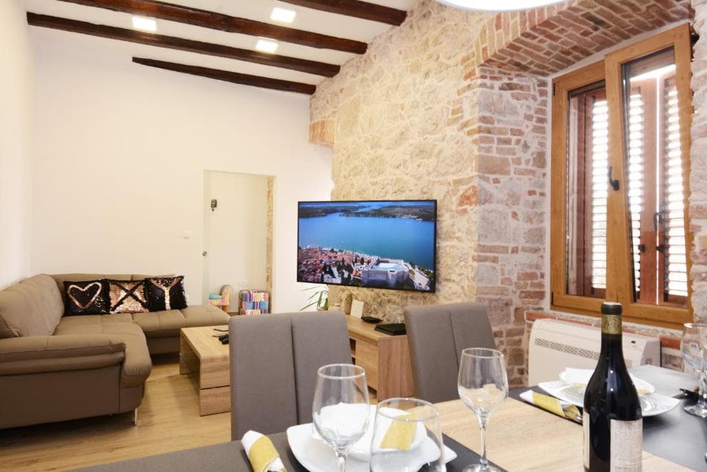 Living and dining area in Design Apartment X with exposed wooden beams, a stone wall with a shuttered window, a sofa, a TV and a set dining table