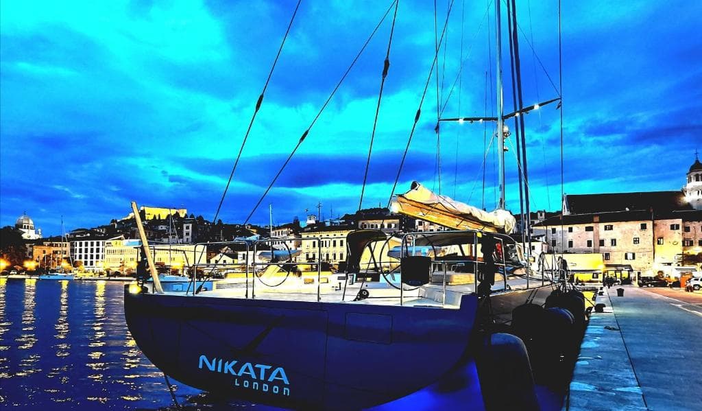 Sailing yacht moored on the Šibenik waterfront at dusk, with the old town lit up behind